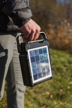 Bir güneş panelinin dikey fotoğrafı. Bir adam, doğanın arka planına karşı sağ elinde bir şarj istasyonu taşır. Şarj istasyonu batarya ve güneş paneli ile