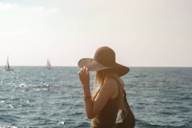 Siyah şapkalı bir kız denize doğru bakıyor. Ufukta iki yelkenli var. Model, deniz kıyısında güneşli havada bir şapkayı buduyor. Çerçeve güneş tarafından aydınlatılır.