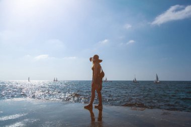 Model, arka ışıklandırılmış güneş ışığında deniz kenarında bir rıhtımda duruyor. Kız yelkenlilerdeki yatlarda denize doğru bakıyor. Deniz kenarında dinlen. Kız şort, tişört ve şapka giymiş..