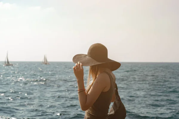 A girl in a black hat looks towards the sea. Two sailboats are sailing on the horizon. The model trims a hat in sunny weather on the seashore. The frame is illuminated by the sun