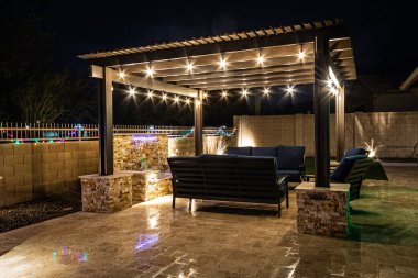 A resort style backyard at night with a waterfall, pergola, and a firepit at night.