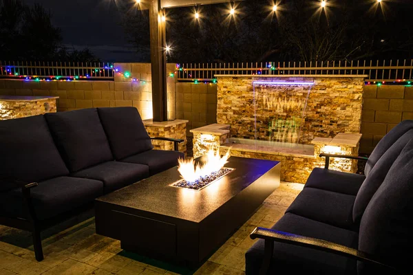 A resort style backyard at night with a waterfall, pergola, and a firepit at night.