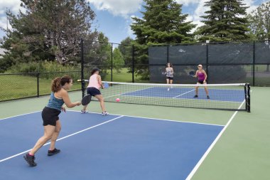 A comptetivie doubles game of pickleball with a group of women on a blue and green court in summer.