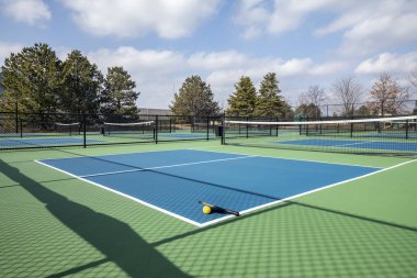 View of a pickleball complex with a paddle and yellow on blue and green courts beside a playground in a suburban park in early spring. clipart