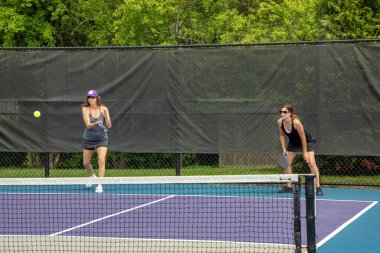A pickleball player serves while her partner prepares for a return on a suburban pickleball court during summer.