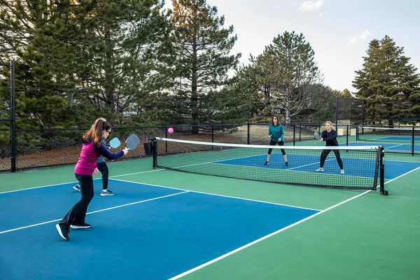 Four women pickleball players volley a pink ball near the net on a blue and green court in early spring.