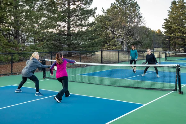 Four women pickleball players volley near the net on a blue and green court in early spring.
