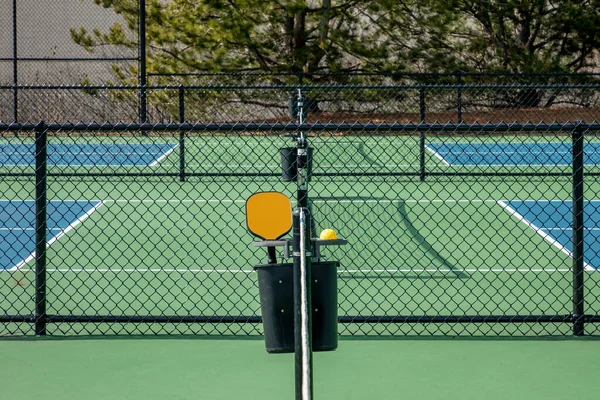 郊外の複合施設で黒フェンシングと青と緑の裁判所の間のオレンジ色のピックルボールパドルと黄色のボールの眺め ストック画像