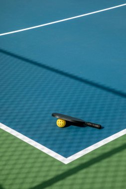 Pickleball raketi ve sarı top beyaz çizgiler ve net gölgeli mavi ve yeşil bir kortta.