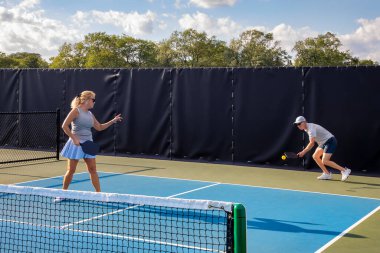 Erkek bir picleball oyuncusu, kadın partneri topun içeride olup olmadığını kontrol ederken topu geri verir..