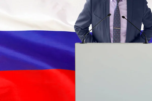 ロシアの国旗を背景にマイクと男とのトリビューン ビジネスマンとロシアの旗の背景にトライブリーン マイクの背景を持つ表彰台の政治家ロシアの旗 — ストック写真