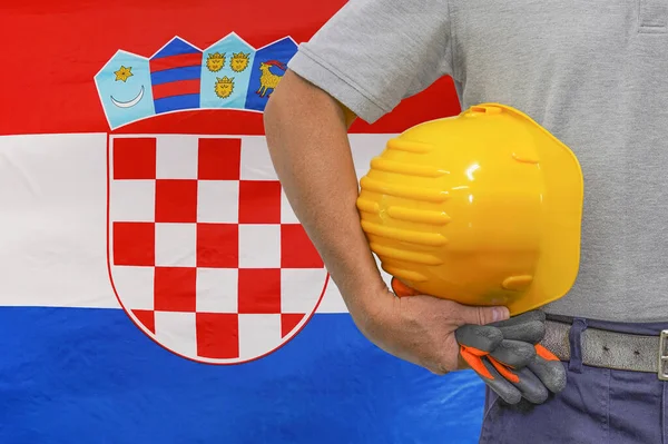 Close-up of hard hat holding by construction worker on flag of Croatia background. Hand of worker with yellow hard hat and gloves. Concept of Industry, construction and industrial workers in Croatia