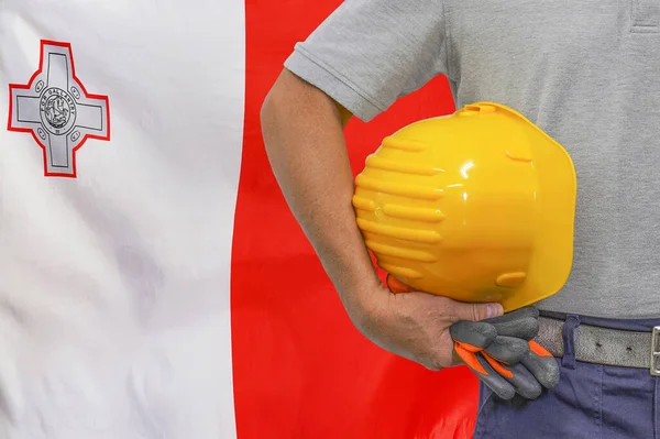 Close-up of hard hat holding by construction worker on flag of Malta background. Hand of worker with yellow hard hat and gloves. Concept of Industry, construction and industrial workers in Malta