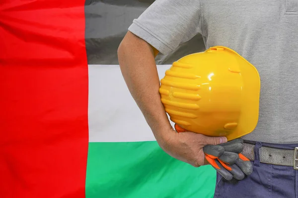 Close-up of hard hat holding by construction worker on flag of United Arab Emirates background. Hand of worker with yellow hard hat and gloves. Concept of industrial workers in United Arab Emirates