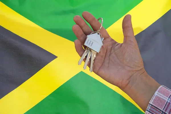 Hand shows the keys to the apartment on background of flag Jamaica. Keys to the apartment in a female hand on background of flag Jamaica. Realty concept and home buying or new apartment in Jamaica