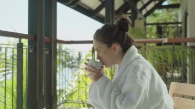 Beyaz otel bornozlu genç kadınlar tropikal tatil beldelerinde balkonda içmeyi severler. Tatil modu ve rahatlatıcı spa oteli. Sıcak bir günde arka planda palmiye ağaçları olan tatilde ya da izinli bir kadın