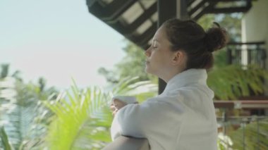 Tropikal plaj tatil beldesindeki kadınların balkonunda tatilin tadını çıkarıyor ve güneşleniyor. Lüks spa otelinde harika bir tatil. Beyaz bornozla gülümseyerek ve nefes alarak kahve ya da çay içerek.