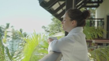 Otel beyaz cüppeli genç kadın tropikal tatil köyündeki bardaktan nefes alıp içiyor. Spa otelinin balkonunda dikilip tatilin ya da tatilin tadını çıkarıyorum. Arka planda palmiye ağaçları var. Güneşin altında dinleniyor