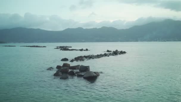 在非洲热带岛屿旁边的下午阳光下 海水中的岩石和石头 夕阳西下 平静的海水和波浪冲向岸边 遥远的主岛和城市背景 非洲的塞舌尔 — 图库视频影像