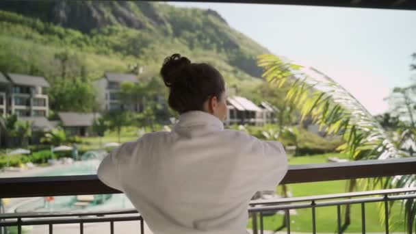 白いローブを着た若い女性がスパホテルからの熱帯の島の景色を楽しんでいます 美しい景色がリラックスして穏やかな時間で豪華な休暇や休日 プールを見下ろすバルコニーの女性 — ストック動画
