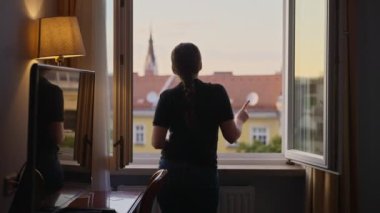 Otelde ya da apartmanın penceresinde durup dans eden ve anın tadını çıkaran bir kadın. Seyahat sırasında ya da tatilde bir turist ya da genç bir bayan gezgin odada eğlenceli ve enerjik bir şekilde dans ediyor.