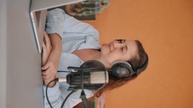 Dikey video - video konferansında kadın sunucu ya da konuk ya da görüşme. Genç bir kadın stüdyosunda ya da ofisinde dizüstü bilgisayarına bakıyor ve bir hoparlörü dinliyor. Başını salla ve gülümse.