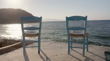 Yunanistan 'da öğleden sonra güneşinde uçurum ve denizin önüne iki açık mavi sandalye kurulmuş. Akdeniz 'e bakan mavi renkli klasik taverna sandalyeleri. Dalgalar çarpıyor ve güneş parlıyor.