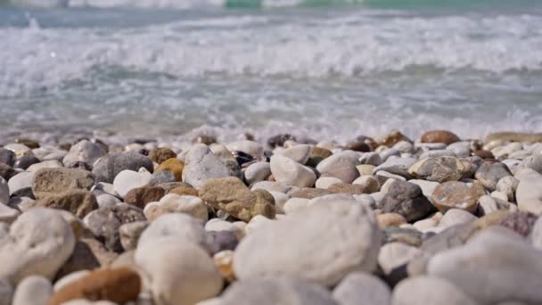 海浪来袭时 海滩或海岸上的岩石和小河石 蓝色的海浪在背景上飞溅 炎热而阳光灿烂的夏日 地上布满了五彩缤纷的石头和岩石 靠近点 — 图库视频影像