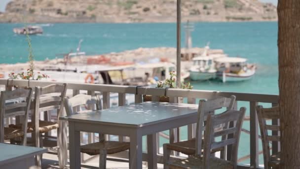 地中海の日当たりの良いクレタ島のギリシャの島で美しく 平和なタヴェルナ 背景にドックや漁船と青い澄んだ水 伝統的なギリシャ料理レストランの椅子とテーブル — ストック動画
