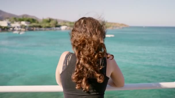 風が吹くと若い女性旅行者は休暇中に青い海を見ている ギリシャの休日女性はギリシャの島でボードウォークのバルコニーの横に立っており ロマンチックな典型的なビューを観察します — ストック動画