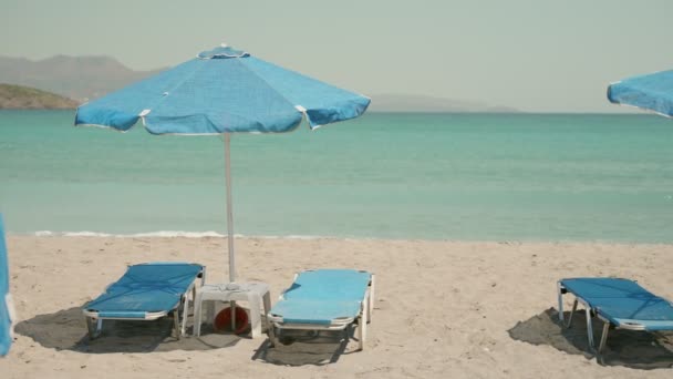 在希腊岛上度假 夏天有海滩的日光浴床和遮阳伞 炎热的天气在沙滩度假胜地与绿松石色海水与海浪冲到岸上 蓝色塑料椅子 — 图库视频影像