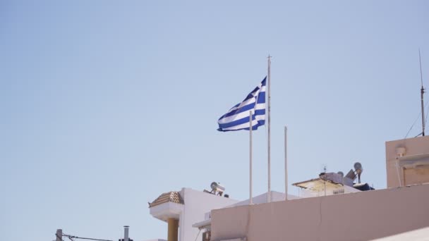 希腊国旗缓缓飘扬 飘扬在附属于一根杆子的建筑物上 希腊一个渔镇的一座建筑物上 希腊蓝白相间的旗帜横扫其间 — 图库视频影像