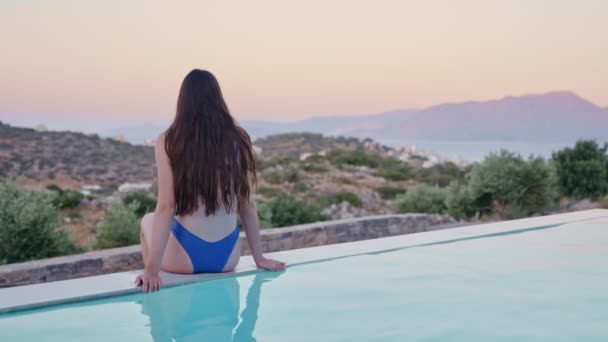 ビキニ水着の若い女性がビューで豪華な家のプールの横に座っています 夕日や日の出で壮大なロマンチックな景色を見下ろすファンシースイートまたはホテルのスイミングプール 女性観光客は地平線を見る — ストック動画
