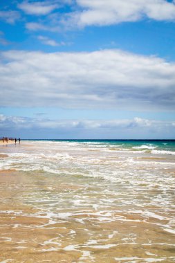 People walk on the wide fine flat sandy beach of Jandia on the left side of the picture, in the south of Fuerteventura, Canary Islands, Spain, vertical format