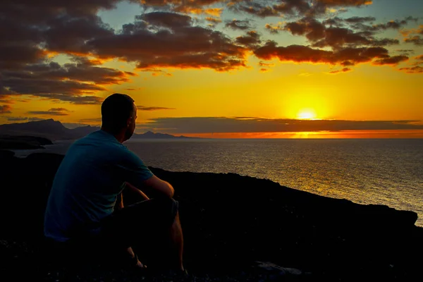 Man sitting on the rocks on the west coast of Fuerteventura at sunset in the evening light and looking out to sea at la Pared, canary islands, spain