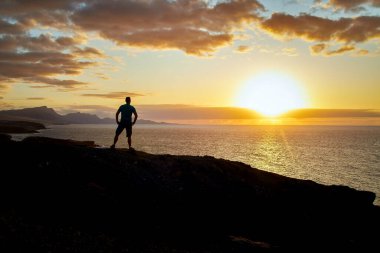 Kanarya Adaları 'ndaki denize ve günbatımına bakan adam akşam ışığında, arka planda La Pared kıyısındaki Fuerteventura dağlarına bakar.