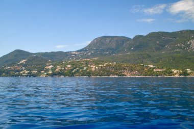 Korfu 'nun doğu sahili, deniz kıyısında dağ yamaçlarında yeşil ormanlar, deniz kıyısında mavi sular ve binalar.