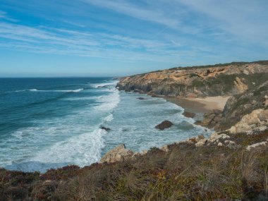 Portekiz, Vila Nova de Milfontes yakınlarındaki yabani Rota Vicentina sahilinde okyanus dalgaları ve keskin kaya ve yeşil bitki örtüsüyle Praia da Angra da Barrela altın kumsal manzarası