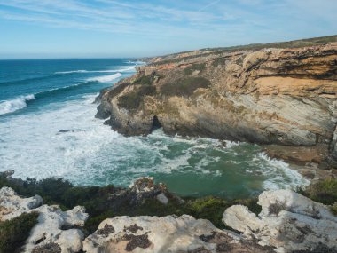 Portekiz, Vila Nova de Milfontes yakınlarındaki Rota Vicentina 'da deniz dalgaları, keskin kayalar ve taşlarla ve yeşil çalılarla kıyı manzarası