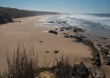 Praia do Brejo Largo sahilinin havadan görünüşü. Okyanus dalgaları, keskin taşlar, kayalıklar ve Rota Vicentina sahilindeki kum. Vila Nova de Milfontes, Portekiz. Güneşli gün, mavi gökyüzü