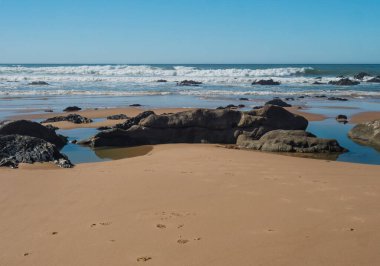Portekiz, Vila Nova de Milfontes yakınlarındaki Rota Vicentina sahilinde okyanus dalgaları ve keskin kayalarla boş Praia do Queimado plajı manzarası.