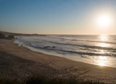 Praia Grande de Almograve kumsalında uzak siluet insanlar ve okyanus dalgaları altın saat ışığı, açık mavi gökyüzü. Rota Vicentina Sahili, Almograve, Portekiz.