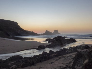 Gün batımından sonra Praia da Zambujeira do Mar in rock and cliff, little stream ve bulanık okyanus dalgaları pembe ışıkta. Rota Vicentina Sahili, Odemira, Portekiz