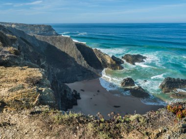 Okyanus dalgaları, uçurumlar ve taşlarla dolu boş Praia das Bernosas plajı, vahşi Rota Vicentina sahillerinde ıslak altın kum ve yeşil bitki örtüsü, Odemira, Portekiz
