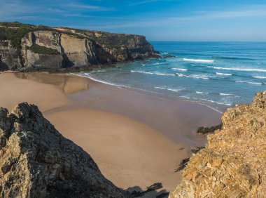 Okyanus dalgaları, uçurumlar ve taşlarla dolu boş Praia do Carvalhal plajı, vahşi Rota Vicentina sahillerinde ıslak altın kum ve yeşil bitki örtüsü, Odemira, Portekiz.