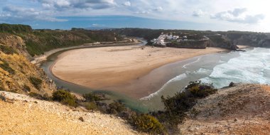 Praia de Aldatmaca 'nın panoramik manzarası Mar Sörfçü plajı altın kum, Atlantik okyanus dalgaları, nehir kıvrımı ve Aldatmaca Köyü' nün beyaz evleri. Rota Vicentina Sahili, Odemira, Portekiz