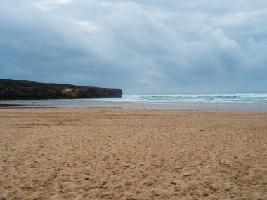 Okyanus dalgaları, uçurum, taşlar ve altın kumun olduğu boş Praia da Amoreira plajı manzarası. Rota Vicentina Sahili, Odemira, Portekiz. Bulutlu karamsar sonbahar günü
