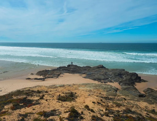 stock image View of Praia dos Aivados sand beach with ocean waves and sharp rock and distant figures at wild Rota Vicentina coast near Porto Covo, Portugal