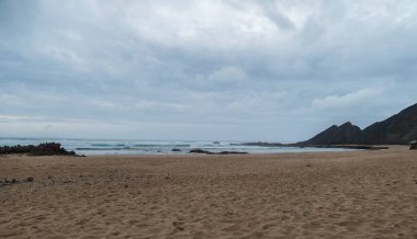 Okyanus dalgaları, uçurum, taşlar ve altın kumun olduğu boş Praia da Amoreira plajı manzarası. Rota Vicentina Sahili, Odemira, Portekiz. Bulutlu karamsar sonbahar günü