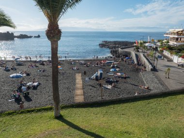 Santiago del Teide, Tenerife, Kanarya Adaları, 18 Aralık 2021: Güneşlenen ve yüzen turist ve palmiye ağaçlarıyla siyah kumsal Playa de la Arena manzarası